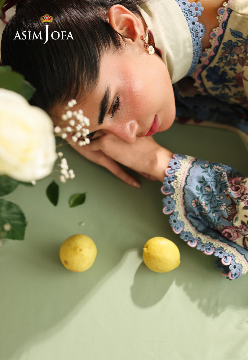 Asim Jofa AJUBC-07 printed lawn campaign portrait – model resting on a table, with blue floral cuff beside yellow lemons